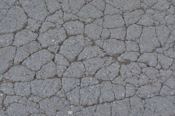 Rocky grey asphalt texture, with small deep cracks and small dark rocks visible in its surface.
